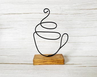 May include: A black wire coffee cup sculpture on a stained wooden base. The cup design is a continuous line, with a handle and steam detail. The sculpture is a decorative item for a kitchen or coffee-themed space.