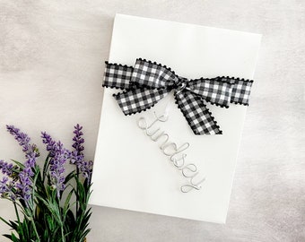 May include: A rectangular white gift box adorned with a black and white gingham ribbon tied in a bow. A silver wire script name tag reading Lindsey is attached to the ribbon. A cluster of purple lavender stems is positioned next to the gift box.