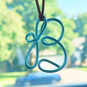 May include: A bright turquoise wire pendant shaped into a cursive letter B, suspended from a thin brown leather cord. The wire is smooth and metallic, forming a continuous loop that creates the stylized character with rounded, flowing lines.