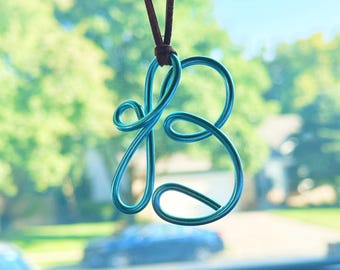 May include: A bright turquoise wire pendant shaped into a cursive letter B, suspended from a thin brown leather cord. The wire is smooth and metallic, forming a continuous loop that creates the stylized character with rounded, flowing lines.