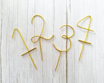 May include: Gold-colored wire cake toppers shaped as the numbers 1, 2, 3, and 4. Each number has a thin, elegant design with a stake at the bottom for insertion into a cake. The toppers are displayed on a light wood-grain surface.