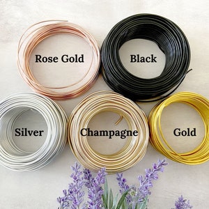 May include: Six spools of wire in different colors: rose gold, black, silver, champagne, gold. The spools are arranged in a circle on a white surface.