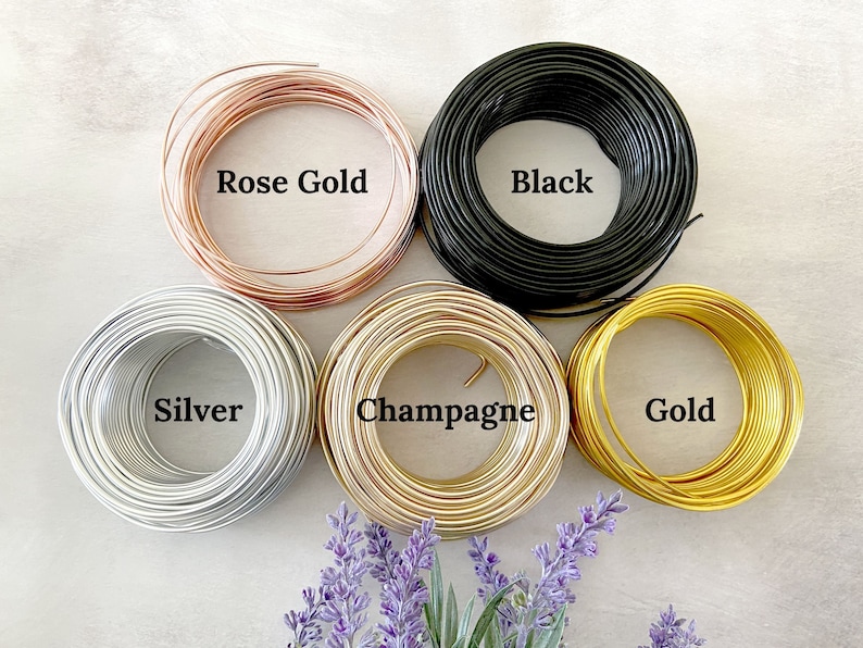 May include: Six spools of wire in different colors: rose gold, black, silver, champagne, gold. The spools are arranged in a circle on a white surface.