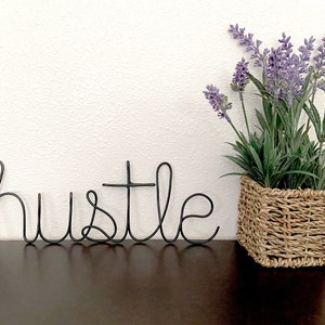May include: Black wire word art spelling "hustle" on a dark brown surface with a potted lavender plant in a woven basket to the right.
