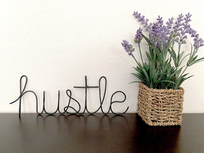 May include: Black wire word art spelling "hustle" on a dark brown surface with a potted lavender plant in a woven basket to the right.