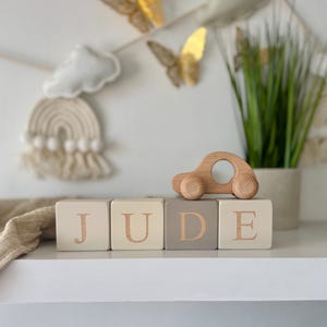 May include: Four wooden blocks arranged in a row, spelling the name JUDE in light brown capital letters. The blocks are painted in shades of cream, beige, and taupe. A small, natural-toned wooden toy car sits on top of the last two blocks.