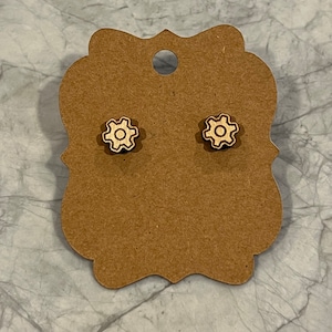 May include: A pair of wooden earrings with a gear design. The earrings are a light brown color with a white gear design.