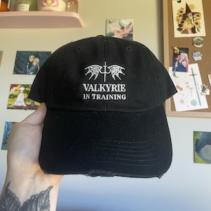 May include: Black baseball cap with a white embroidered design of wings and a sword with the text "Valkyrie in Training".