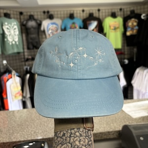 May include: A light blue baseball cap with embroidered celestial design. The cap features a curved brim and a six-panel construction. The embroidery includes stars and a crescent moon. The cap is held up by a person's arm.
