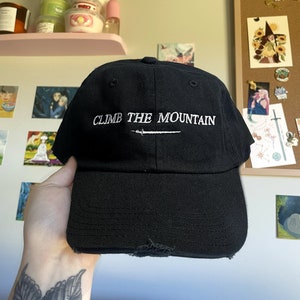 May include: Black baseball cap with a white embroidered design that says "CLIMB THE MOUNTAIN" with a small sword graphic underneath the text.
