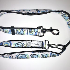 Puede incluir: Un juego de correa y asa para perros. La correa y el asa presentan un fondo blanco con un estampado floral azul, azul claro y amarillo. Los bordes son negros y los herrajes plateados.