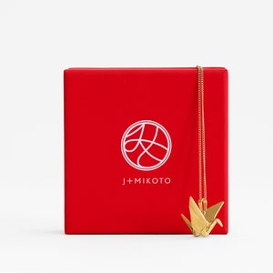Peut inclure: Une bo&icirc;te rouge avec un logo blanc qui dit "J+MIKOTO" et un cercle blanc avec un design stylis&eacute; &agrave; l'int&eacute;rieur. Un collier en or en forme de grue origami est suspendu &agrave; la bo&icirc;te.