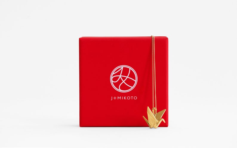 Peut inclure: Une bo&icirc;te rouge avec un logo blanc qui dit "J+MIKOTO" et un cercle blanc avec un design stylis&eacute; &agrave; l'int&eacute;rieur. Un collier en or en forme de grue origami est suspendu &agrave; la bo&icirc;te.