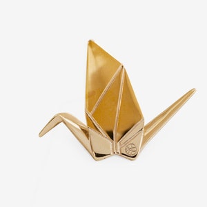 Peut inclure: Une sculpture de grue en origami dor&eacute;e. La grue est pli&eacute;e dans un style traditionnel et a un petit cercle avec un motif au centre de sa poitrine.