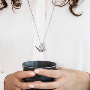 Peut inclure: Un collier en cha&icirc;ne argent avec un pendentif en origami de grue argent&eacute;. Le collier est port&eacute; par une personne tenant une tasse en c&eacute;ramique gris fonc&eacute;.