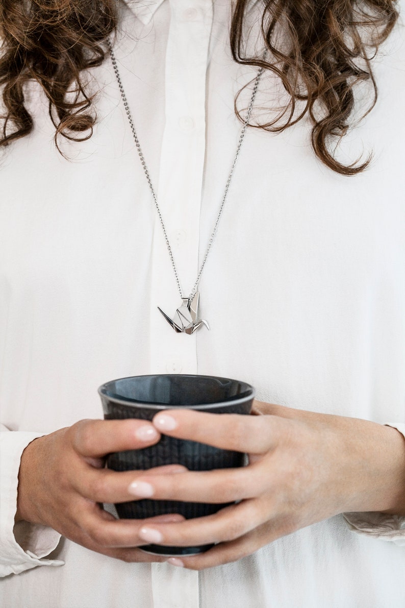Peut inclure: Un collier en cha&icirc;ne argent avec un pendentif en origami de grue argent&eacute;. Le collier est port&eacute; par une personne tenant une tasse en c&eacute;ramique gris fonc&eacute;.