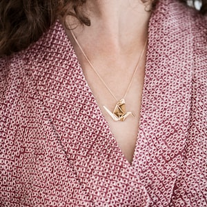 Peut inclure: Un collier avec un pendentif en forme de grue origami en or sur une cha&icirc;ne d&eacute;licate. Le collier est port&eacute; par une personne aux cheveux bruns boucl&eacute;s et portant une chemise &agrave; motifs roses.