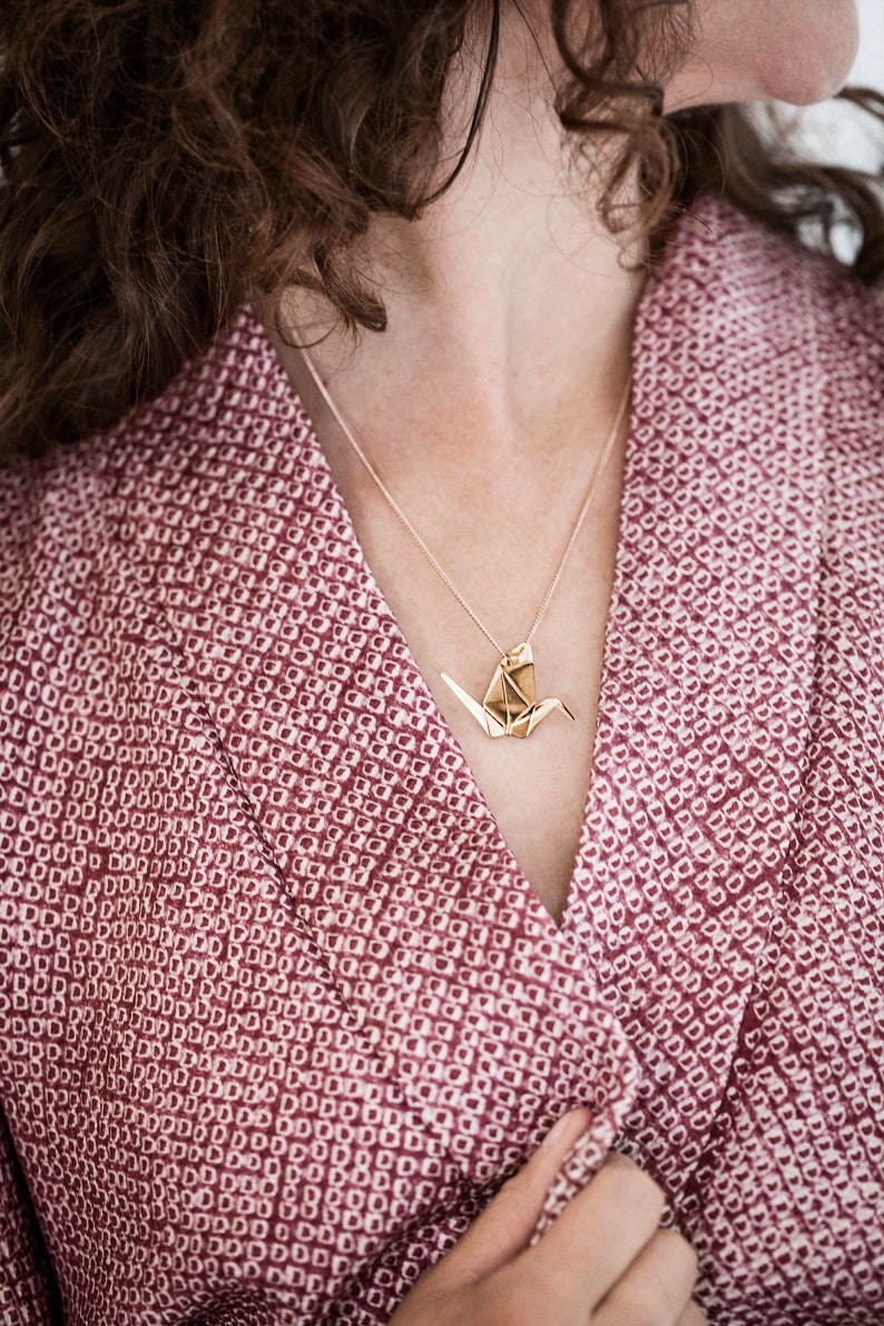 Peut inclure: Un collier avec un pendentif en forme de grue origami en or sur une cha&icirc;ne d&eacute;licate. Le collier est port&eacute; par une personne aux cheveux bruns boucl&eacute;s et portant une chemise &agrave; motifs roses.