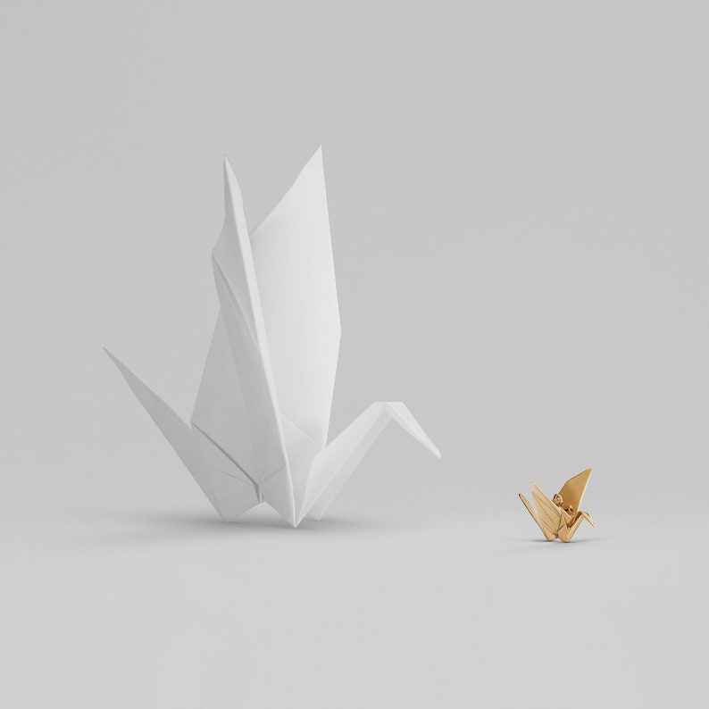 Peut inclure: Une grue en origami blanche se tient debout, face &agrave; une grue en origami dor&eacute;e plus petite. Les deux grues sont pli&eacute;es en papier et sont positionn&eacute;es sur un fond blanc.