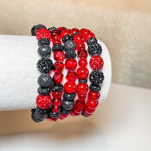 May include: A stack of six bracelets with red and black beads. The bracelets are made of different materials, including lava rock, glass, and crystal beads.