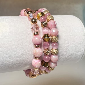 May include: Three pink and gold beaded bracelets. The bracelets are made with pink and white marbled beads, gold beads, and clear crystal beads.