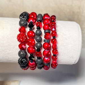 Könnte beinhalten: Ein Set aus sechs roten und schwarzen Perlenarmbändern. Die Armbänder bestehen aus roten Glasperlen, schwarzen Lavasteinen und schwarzen Kristallperlen. Die Armbänder sind übereinander gestapelt.