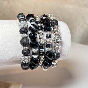 Op de afbeelding: Een set van zes armbanden met zwarte lavasteen, zwarte gefacetteerde glazen kralen, zilveren metalen kralen en zilveren spacer kralen. De armbanden zijn gestapeld op een wit oppervlak.