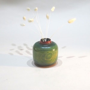 Pode incluir: Um pequeno vaso de madeira redondo com acabamento verde e amarelo. O vaso tem uma borda vermelha e contém vários caules de flores brancas secas e fofas. O vaso está sobre uma superfície clara.