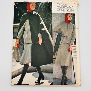 May include: Vintage Vogue Americana Anne Klein fashion magazine cover. The cover shows a woman in a gray suit and a dark green cape. The woman wears a red scarf and holds a cane. The magazine is set against a white backdrop.