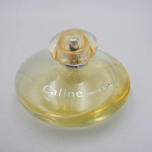 May include: A clear glass bottle of perfume with a gold cap. The bottle is round and has a textured surface. The label on the bottle reads "Caline Parfums Cr".