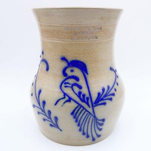 May include: A tan stoneware vase featuring a cobalt blue hand-painted bird and floral motif. The upper portion is stamped with the text Beaumont Bros. Pottery, S. RUCKER, Crooksville, Ohio. The vessel has a wide, flared rim and a rounded body.