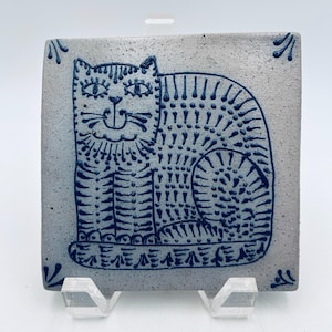 May include: A square ceramic tile with a light gray speckled finish featuring a dark blue hand-painted illustration of a seated cat. The cat design is composed of intricate lines and dots, with small decorative corner accents on the tile surface.