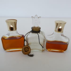 May include: Three vintage perfume bottles with gold caps and clear glass. The center bottle has a clear glass stopper shaped like a flower with an "R" on it.
