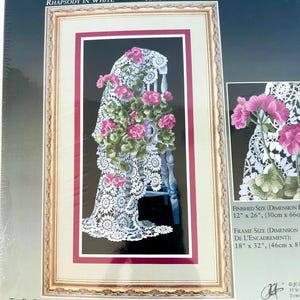 May include: A cross stitch kit titled Rhapsody in White featuring a white lace cloth draped over a chair with pink geranium flowers and green leaves against a black background. The finished size is 12 inches by 26 inches and frame size is 18 by 32 inches.