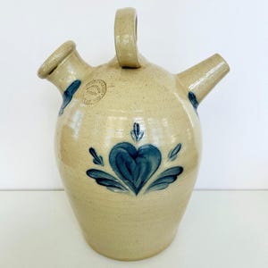 Pode incluir: Um jarro de grés bege com acabamento salpicado, apresentando um motivo floral azul em forma de coração, uma pega superior e dois bicos. Um selo circular na parte superior do corpo contém o texto "STRAPOWITZ POTTERY LAXAN-WEB".