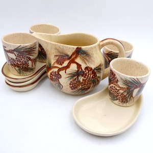 May include: A cream-colored ceramic tea set with a pine cone and branch design. The set includes a pitcher, four cups, and four saucers.