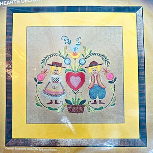 May include: A square embroidery kit titled HEARTS DELIGHT featuring a central red heart, a blue bird, and two figures in traditional clothing. The design is framed by a yellow border and a dark wood-grain frame, with text at the bottom.