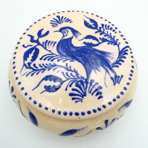 May include: A round, off-white ceramic trinket box with a blue bird and floral design on the lid. The bird is surrounded by blue leaves and vines. The rim of the lid is decorated with small blue dots.