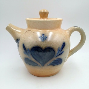 May include: A tan ceramic teapot with a lid and handle. The teapot is decorated with a large blue heart and blue leaf designs. The spout and handle are curved. The lid has a small knob.