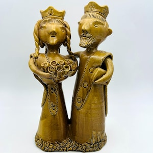 Op de afbeelding: Een keramische sculptuur van een koninklijk paar. De figuren zijn gekleed in sierlijke gewaden en kronen, waarbij de vrouw een boeket vasthoudt. De sculptuur heeft een warme, aardse bruine kleur en een getextureerde afwerking. Het paar staat dicht bij elkaar.