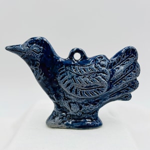 May include: A dark blue ceramic bird ornament with a glossy finish and a speckled texture. The bird features an embossed wing pattern with linear details and a small circular hole at the top for hanging, showcasing a stylized avian silhouette.