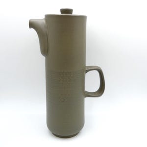 May include: A tall, cylindrical, matte taupe ceramic coffee pot featuring a smooth upper section and a lower section decorated with a repeating, embossed herringbone pattern. It includes a curved handle, a small spout, and a flat lid.