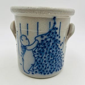May include: A speckled light grey stoneware crock featuring two side handles and a blue hand-painted design of a figure decorating a Christmas tree. The cylindrical vessel has a thick rim and a textured surface with vertical blue lines.