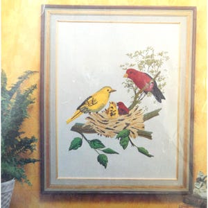 May include: A framed embroidery art piece featuring a yellow bird and a red bird perched on a branch with a nest containing two small chicks. The artwork is set against a light background and enclosed in a simple wooden frame with a light border.