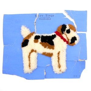 May include: A handmade dog-shaped rug with a red collar, featuring a white, brown, and black pattern. The rug is placed on a torn blue paper with the text "TUFT-TINGS WASHABLE".