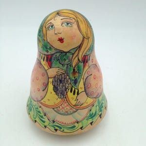 May include: A hand-painted wooden Matryoshka doll, depicting a woman with blonde hair and a green headscarf. The doll holds grapes. The design incorporates pink, yellow, and green, in a traditional folk art style.
