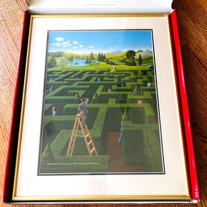 Op de afbeelding: Ingelijste prent van een groen heggenlabyrint met mensen die de paden verkennen. Een man op een ladder kijkt uit over het labyrint. De lucht is blauw met wolken. De prent zit in een gouden lijst en een rode doos.