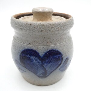 May include: A speckled light gray stoneware crock with a matching lid and a dark blue hand-painted heart design on the front. The ceramic container features a textured, earthy finish with a wide rim and a knobbed lid, showcasing a rustic appearance.
