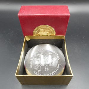 May include: A clear glass paperweight featuring an etched Mars rover design, with the text "Mars Exploration Rovers 2004 NASA/Caltech/JPL". It is presented in a gold-lined box with a red lid.