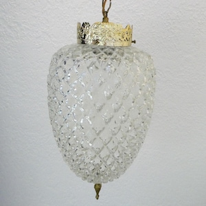 May include: Clear glass pendant light with a diamond pattern. The light fixture has a gold-colored crown-shaped top and a small, decorative bottom piece. The light hangs from a wire and is designed to be a ceiling fixture.